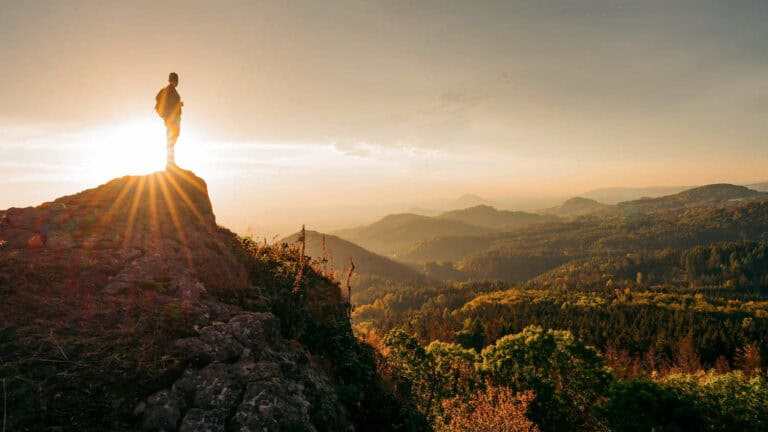 Man in silhoutte standing on top of rocky cliff in front of rolling forested hills